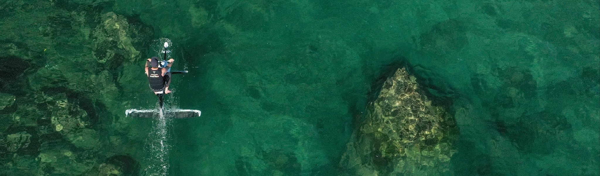 Top view of a male riding a hydrofoiler Xe-1 on clear water ocean showing rocks on the bottom
