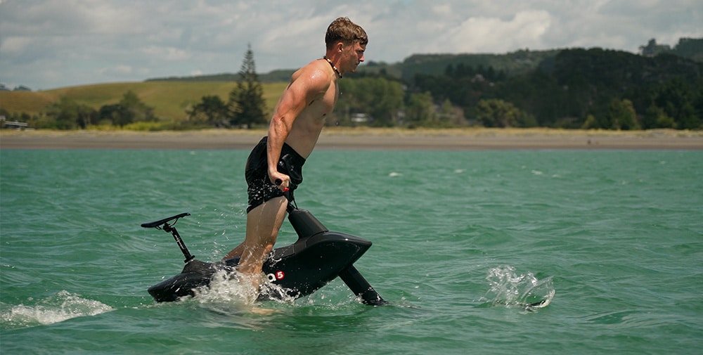 Male performing a deep water start on a black Hydrofoiler SL3 water-bike on the ocean with a beach on the background